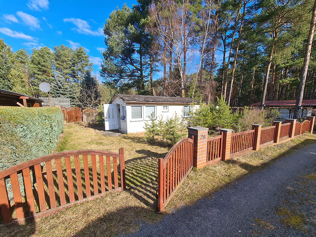 Ansicht eines kleinen weißen Bungalows. Drumherum eine grüne Gartenfläche. Im Hintergrund stehen hohe Bäume.