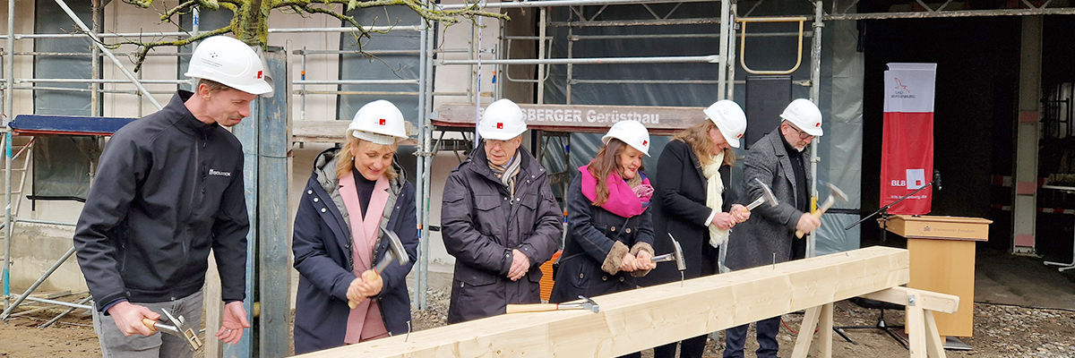 Bild: Personen mit Bauhelm stehen hinter einem Balken und schlagen Nägel ein.