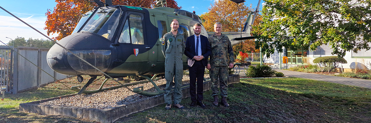 Bild: Zwei uniformierte Soldaten stehen neben einem Mann im Anzug und lächeln in die Kamera. Hinter ihnen stehet ein militär-grüner Helikopter.