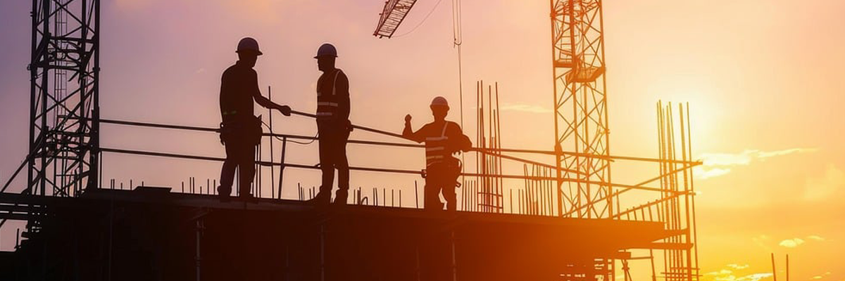 Bild: Bild einer Baustelle mit Sonnenuntergang. Auf einem Gerüst stehen drei Personen. Im Hintergrund sind zwei Baukräne zu sehen.