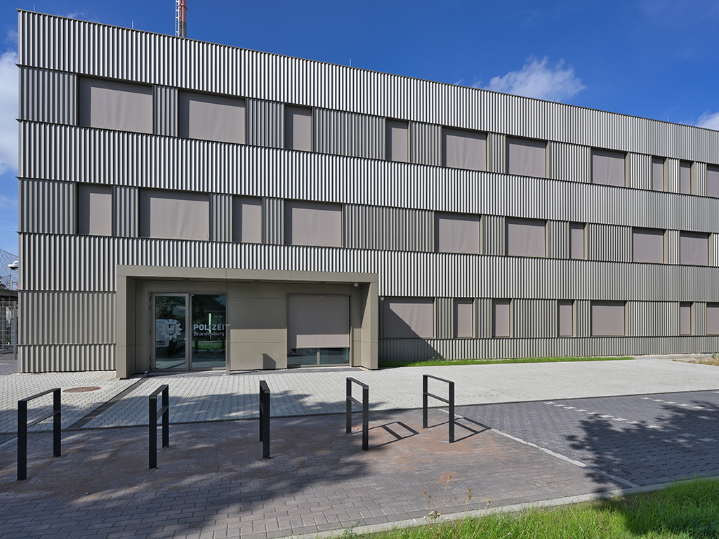 Außenansicht des Gebäudes. Es ist mit grauem Faltblech verkleidet. An der Tür ist das Polizei-Logo und der Name zu sehen. Vor dem Gebäude stehen Fahrragständer.