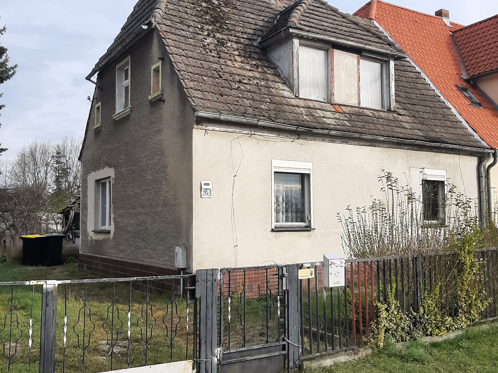 Vorderansicht eines Doppelhauses, linke Seite. Die Fassade ist grau und an der Seite leicht verwittert. Im Dach ist ein Gaubenfenster. Um das Haus ist ein eingezäuntes Grundstück.