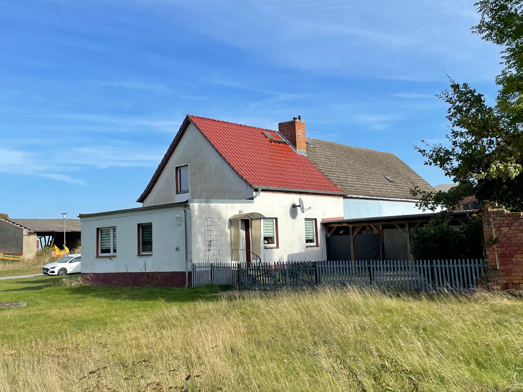 Blick auf die Stirnseite einer Doppelhaushälfte mit Anbau. Die Fassade ist weit, das Dach ist rot geziegelt. Im Garten steht ein Carport. Der Garten ist umzäunt.