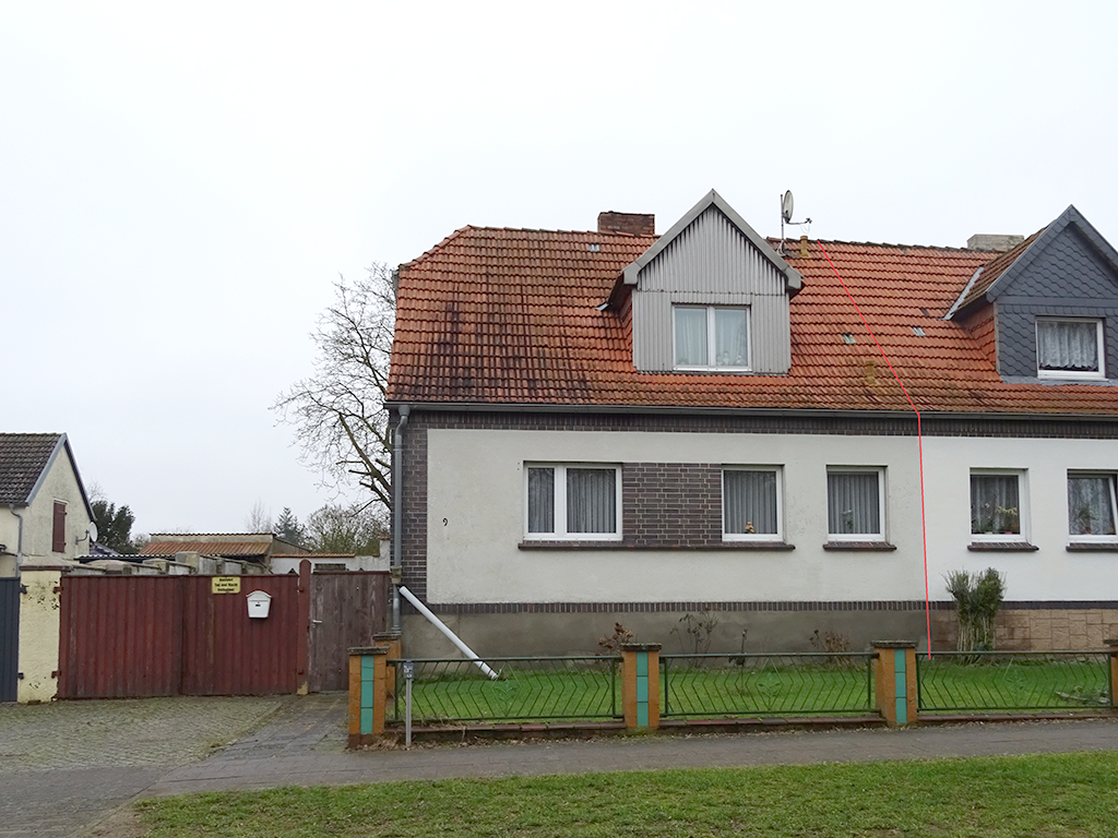 Frontansicht auf einer Doppelhaushälfte (links). Der Putz ist leicht ergraut. Als Kontrast befinden sich Klinkerelemente an der Fassade. Auf dem Dach ist ein großes Gaubenfenster.