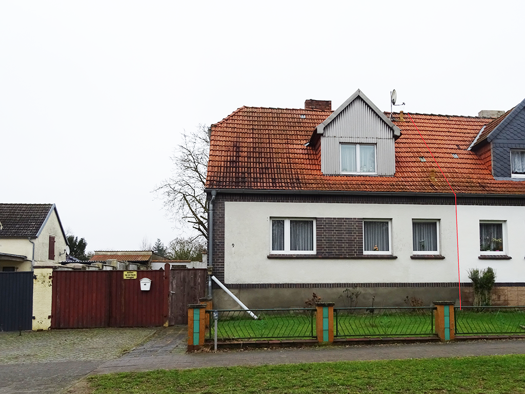 Frontansicht einer Doppelhaushälfte. Markiert ist die linke Seite. Das Haus hat eine weiße Fassade mit Klinkersteinen um die Fenster. Links führt eine Toreinfahr auf den Hof.