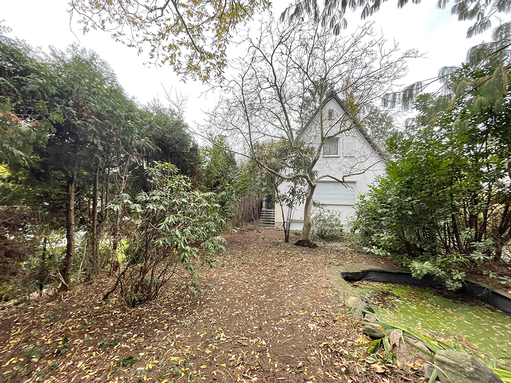 Giebelansicht eines Hauses mit weißer Fassade. Es ist umgeben von einen Grundstück mit viel Baumbestand.