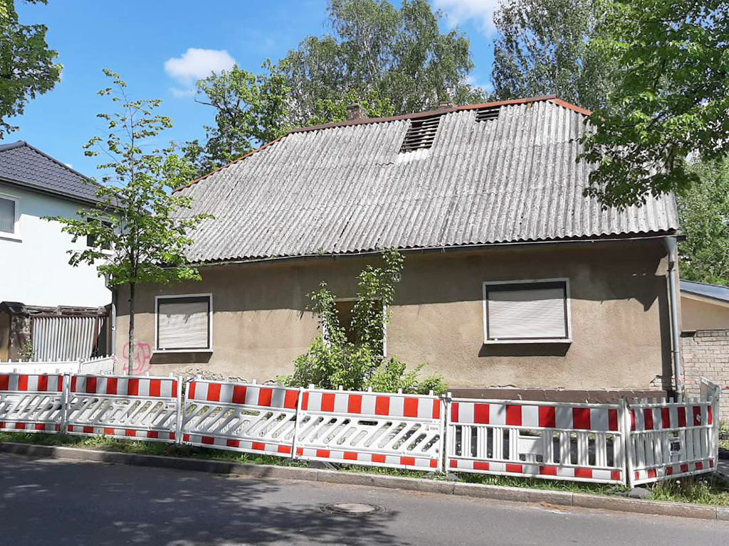 Frontaufnahme eines alten Gebäudes mit geschlossenen Fensterläden. Das Dach ist undicht. Vor dem Haus sind rot-weiße Absperrungen.