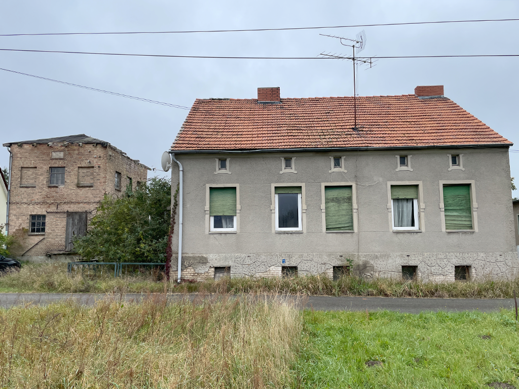 Außenansicht eines alten Hauses: grauer Putz, kaputte Rolläden und Fenster. Daneben steht ein Backsteinhaus mit kaputtem Dach.