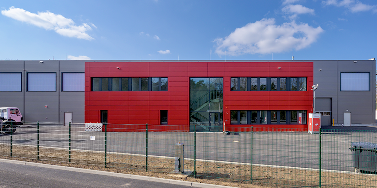 Frontansicht eines zweistöckigen Gebäudes. Der vordere Teil ist kleiner und rot. Der Hallenkomplex dahinter ist grau.
