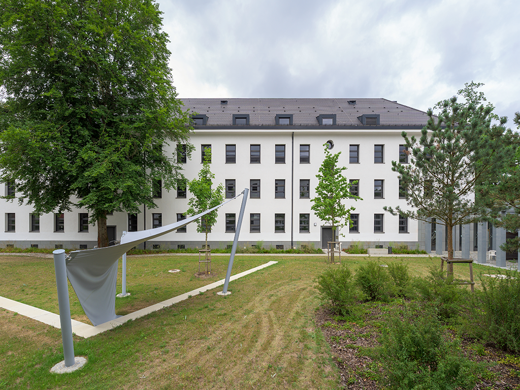 Außenansicht eines dreigeschossigen Gebäudes mit ausgebautem Dach und Grünfläche davor.