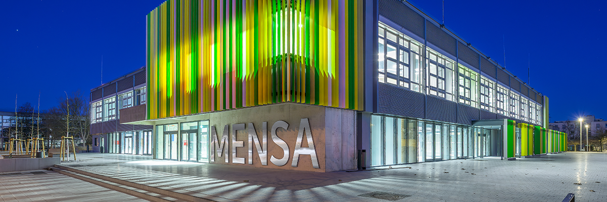 Bild: Außenansicht der Mensa mit auffälligen grünen Lamellen an der Fassade. Das Gebäude ist von Innen beluchtet, der Himmel ist dunkel.