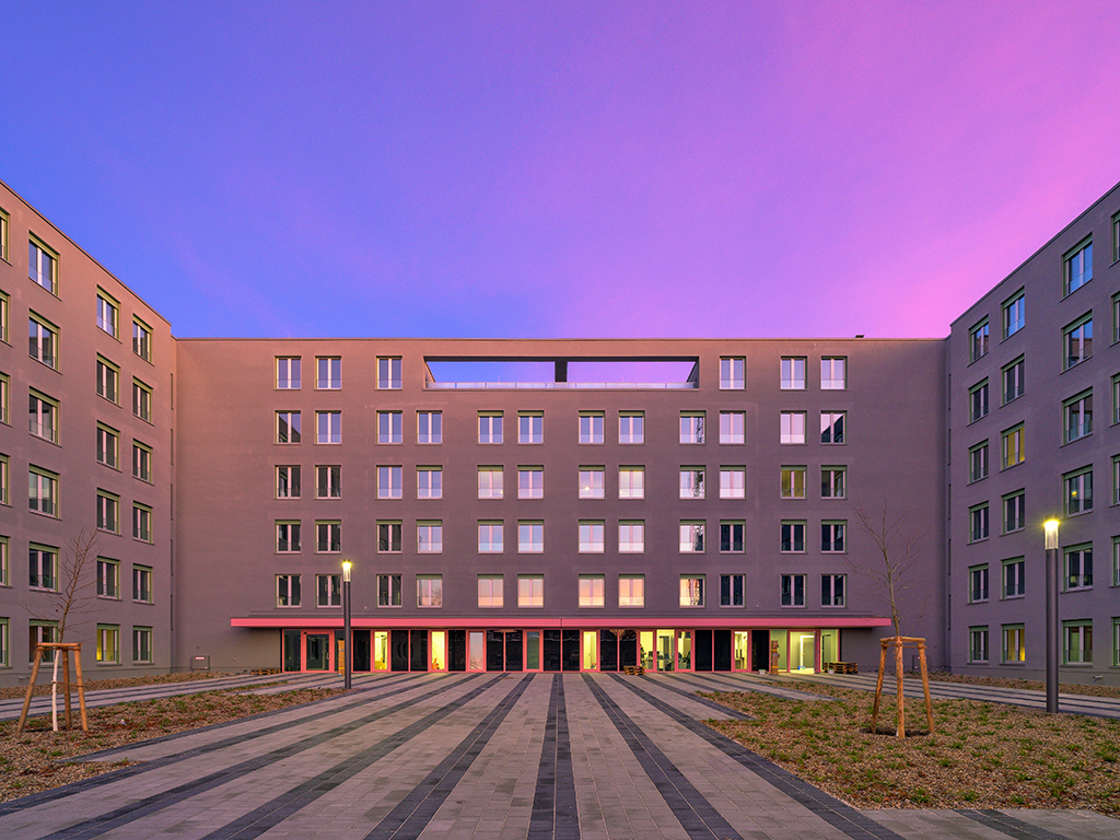 Blick auf einen Platz eines u-förmigen Gebäudes. Der Himmel ist rot gefärbt von der Morgensonne.