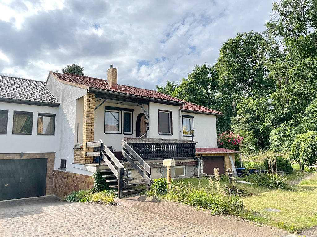 Seitenansicht eines Reihenendhauses mit weißer Fassade. Eine kleine Treppe führt auf eine Veranda zum Eingangsbereich. Links davon geh es abschüssig zur Garage. Vor dem Haus ist ein gestalteter Garten.