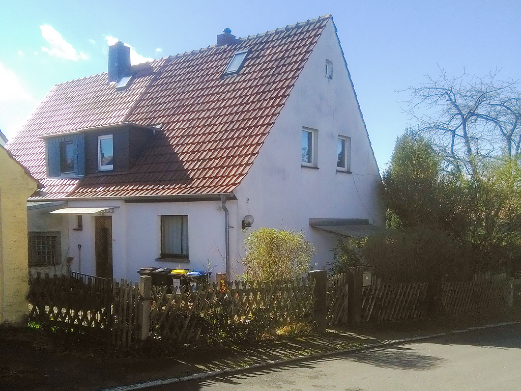 Foto eines Doppelhauses mit zwei Stockwerken und Dachboden, vor einem kleinen Innenhof ist ein niedriger Holzzaun im Vordergrund, im Bild rechts unten ist ein Straße zu sehen