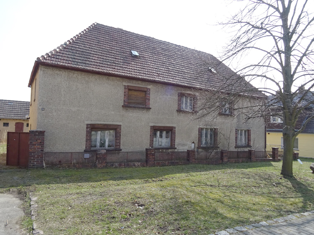 Frontansicht eines altenzweistöckigen Hauses in grauem Rauputz. Davor eine Rasenfläche mit einem Baum.
