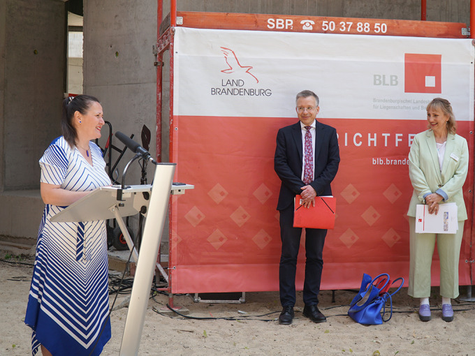 Die Ministerin in einem blau-weißen Kleid hält eine Rede. Im Hintergrund stehen Herr Stolper (StS MdFE) und Gerit Fischer (Technische Geschäftsführerin BLB)