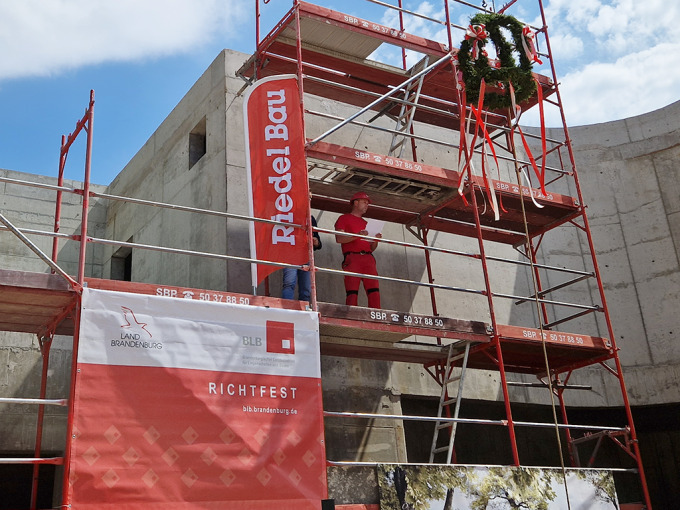Baurbeiter in roter Arbeitskleidung auf einem Baugerüst. Er spricht den traditionellen Richtfestspruch. Oben am Baugerüst ist die Richtkrone gehisst.