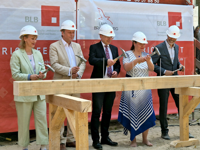 Zwei Frauen und drei Männer stehen hinter einem Balken und schlagen traditionelle den letzten Zimmermannsnagel ein.