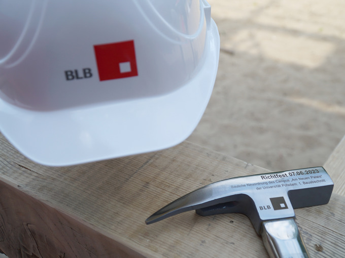 Weißer Bauhelm mit rotem BLB Logo. Davor liegt ein Dachdeckerhammer mit Gravur: Richtfest 07.06.2023. Bauliche Neuordnung des Campus "Neues Palais" der Universität Potsdam: 1. Bauabschnitt.