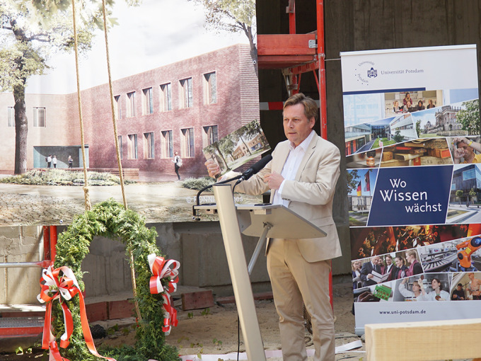 Nahaufnahmen eines Mannes in hellem Anzug. In der Hand hält er ein Architekturbild des Neubaus. Im Hintergrund ist das gleiche Bild auf Bannergröße abgebildet.