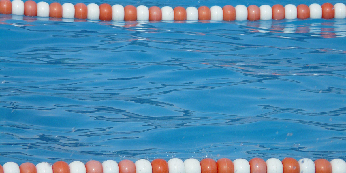 Blaue Wasseroberfläche mit leichten Wellen. Darauf sind im oberen und unteren Bereich des Bildes schwimmende Abtrennungen aus roten und weißen Kugeln.